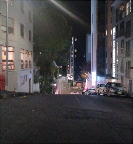 A steep urban street descends between multi-storey buildings, with parked cars, trees, illuminated windows and a junction below.