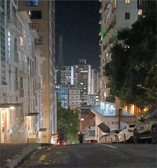 A steep urban street descends between tall buildings, parked cars and trees, with illuminated windows and further buildings in the distance.