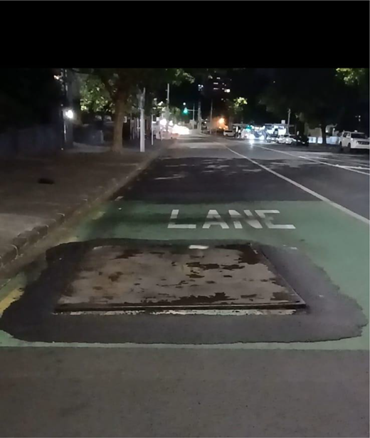 A road lane marked LANE contains a raised rectangular metal plate, with pavements, trees, parked vehicles and traffic lights ahead.