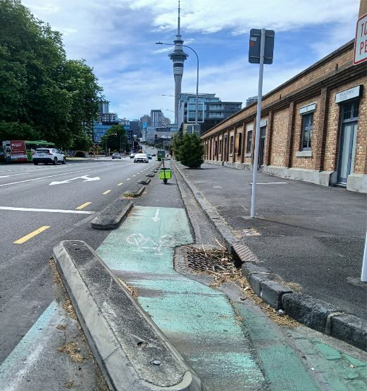 A separated cycle lane runs beside a road and pavement, with raised dividers, drainage grates, vehicles, buildings and a tower in the distance.