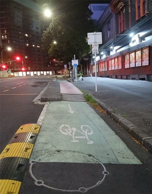 A cycle lane beside a pavement includes bicycle and directional markings, a raised crossing, tactile paving, street signs and traffic lights ahead.