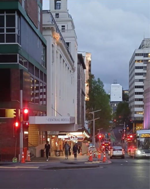 A city intersection contains traffic lights, pedestrians, vehicles, roadworks and tall buildings, including the Central Hotel beside a sloping street.