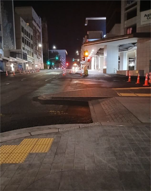 A night-time city street contains tactile paving, a raised kerb, traffic cones, illuminated buildings and traffic lights at roadworks ahead.