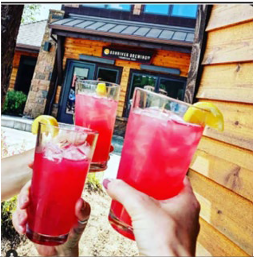 Three hands hold glasses of drinks with lemon slices in front of a wooden building.