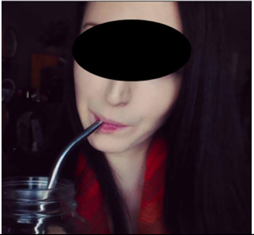 A person with long dark hair drinks from a glass jar using a metal straw, wearing a red and black garment around their shoulders.