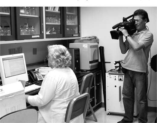 Woman with curly hair seated at a desk and looking at a computer screen next to a phone and boxes, with cabinets, and a man standing on the right holding a large video camera on his shoulder with lab equipment in the background.