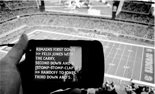 A hand holding a small electronic device with text on its screen describing football game plays and crowd reactions, set against a background of a vast football stadium with a playing field and full spectator stands.
