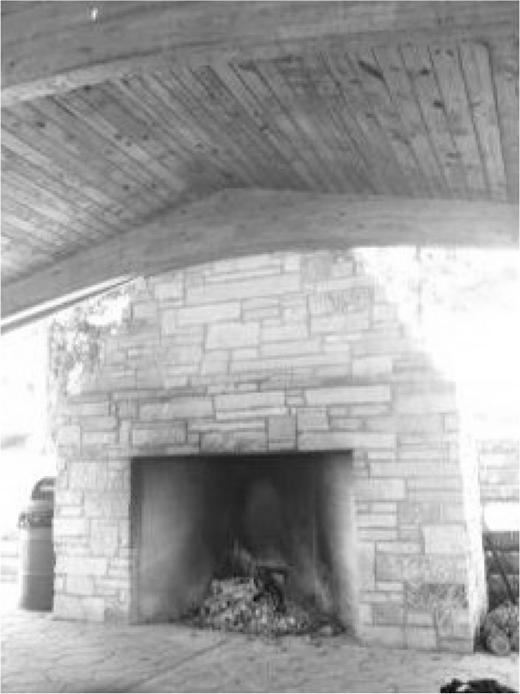 A rustic stone fireplace with a glowing fire within stands under a wooden plank ceiling, flanked by stacked firewood on one side and a barrel on the other.