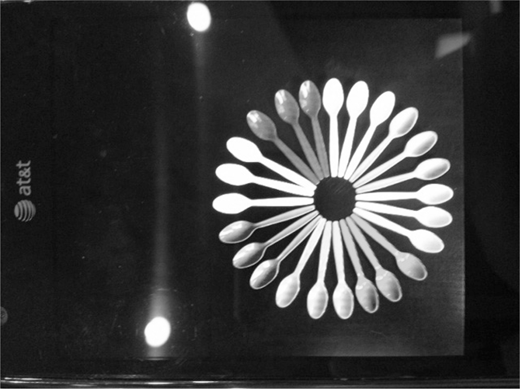 Bright spoons form a circular, sun-shaped arrangement with their handles meeting at a dark center and bowls fanning outward, all resting on a dark surface with a faint A T and T logo along the left side and two glowing oval shapes.