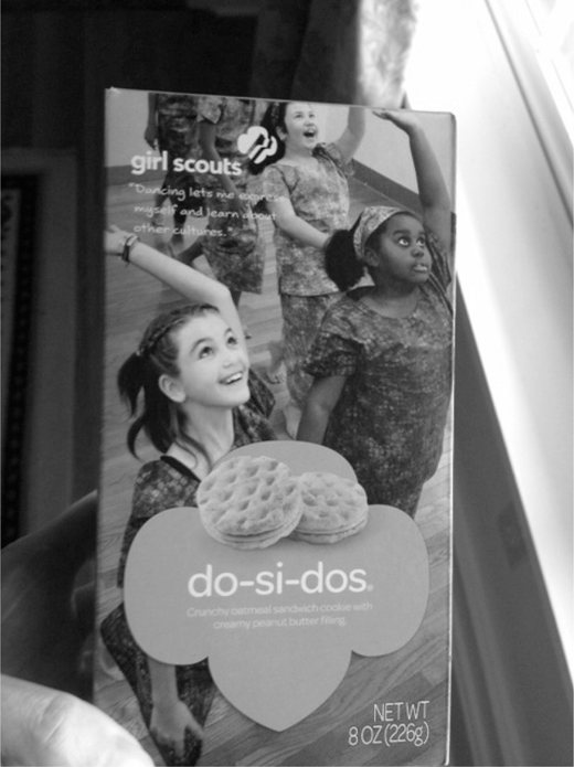 Girl Scouts do-si-dos cookie box, held by a hand, with three young girls in uniforms with raised arms and upward gazes and two round peanut butter sandwich cookies both on the box front.