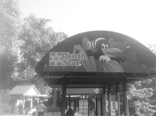 Large arched sign with North Star text and snowflake emblem, along with cartoon marine and bird characters, all above an entrance structure with a metal frame, a backdrop of dense trees, and a small building to the left beside a person.