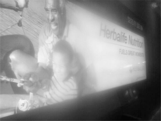 Advertisement on a screen, portraying a joyful older man and a young child together, with the brand name Herbalife Nutrition and the slogan Fuels Great Moments partly discernible through a bright glare on the right.