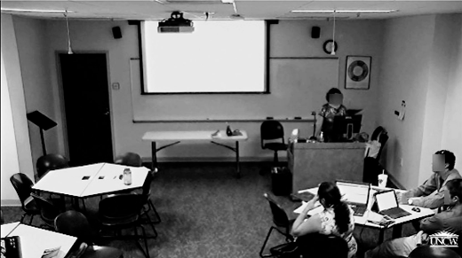 A classroom scene shows a presenter at a podium facing a projection screen, with students seated at tables and working on laptops.