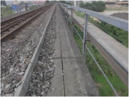 A railway track runs beside a narrow concrete walkway with a metal railing and adjacent road.
