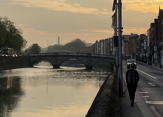 A pedestrian walks along a riverside footpath beside a road and buildings. A multi-arch bridge crosses the river ahead, with a tall obelisk-shaped monument rising in the distant skyline.