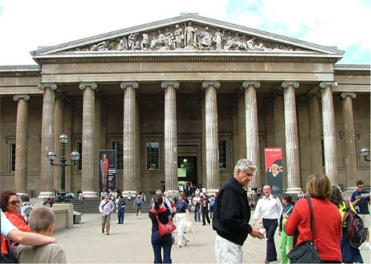 A large classical building with a triangular pediment, sculpted figures, and tall columns. People walk and gather across wide steps and an open forecourt at the main entrance.