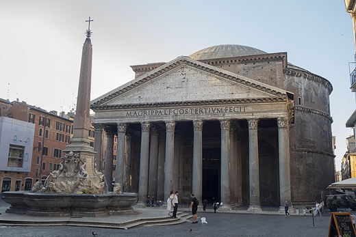 A historic temple with a triangular pediment and tall columns. An inscription appears above the entrance. An obelisk fountain and several people stand in the square.