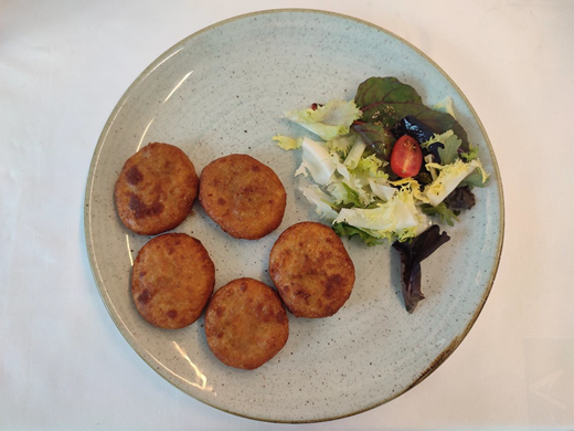 A round plate contains five patties placed on the left side. On the right side, a small portion of mixed salad, including leafy pieces and a halved small tomato is placed.